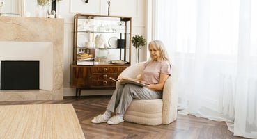 Older woman sitting in an armchair in a well-lit living room, reading a book
