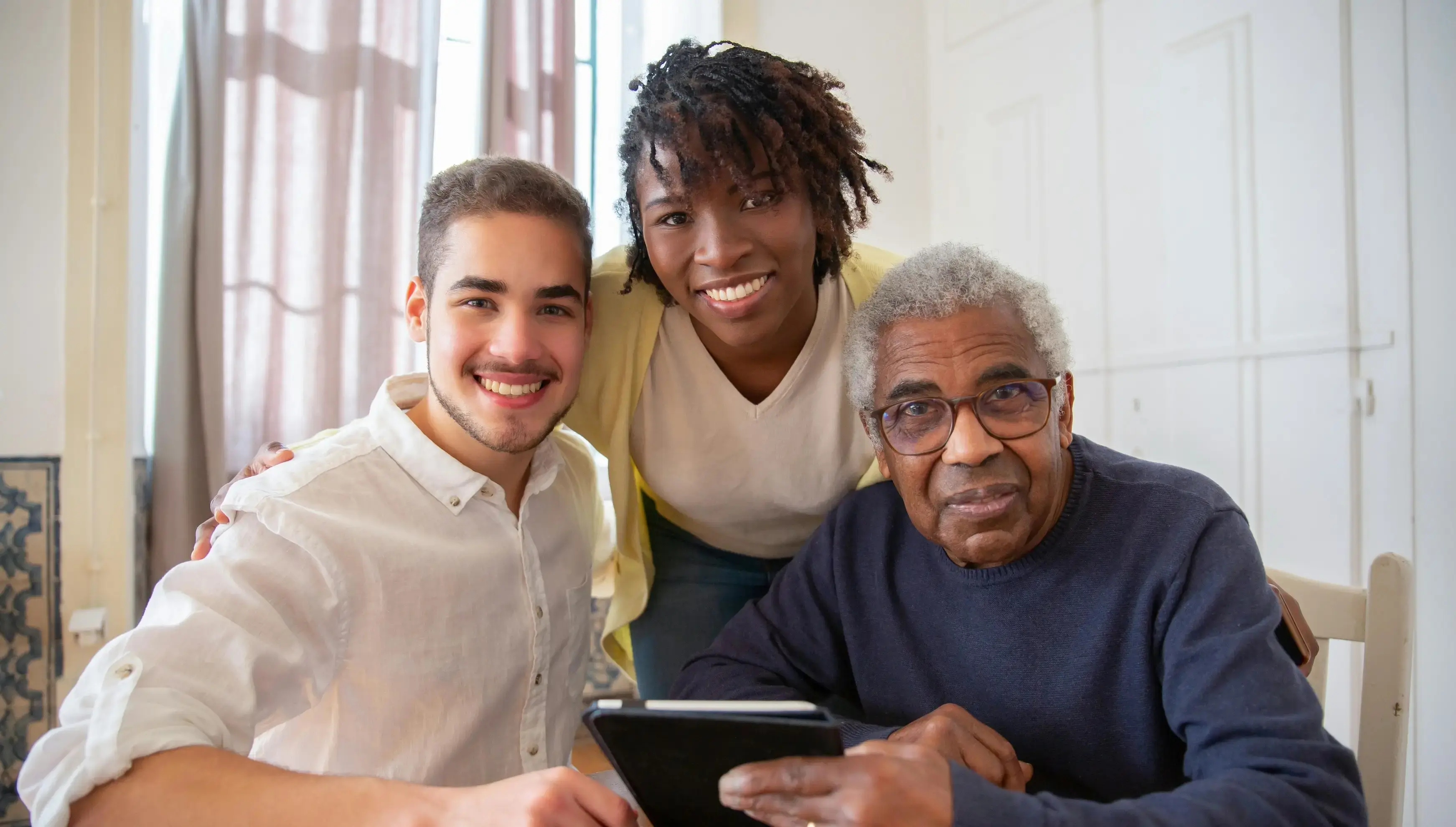 A senior adult man sitting at a table with his two younger caregivers, a young man and a young woman.