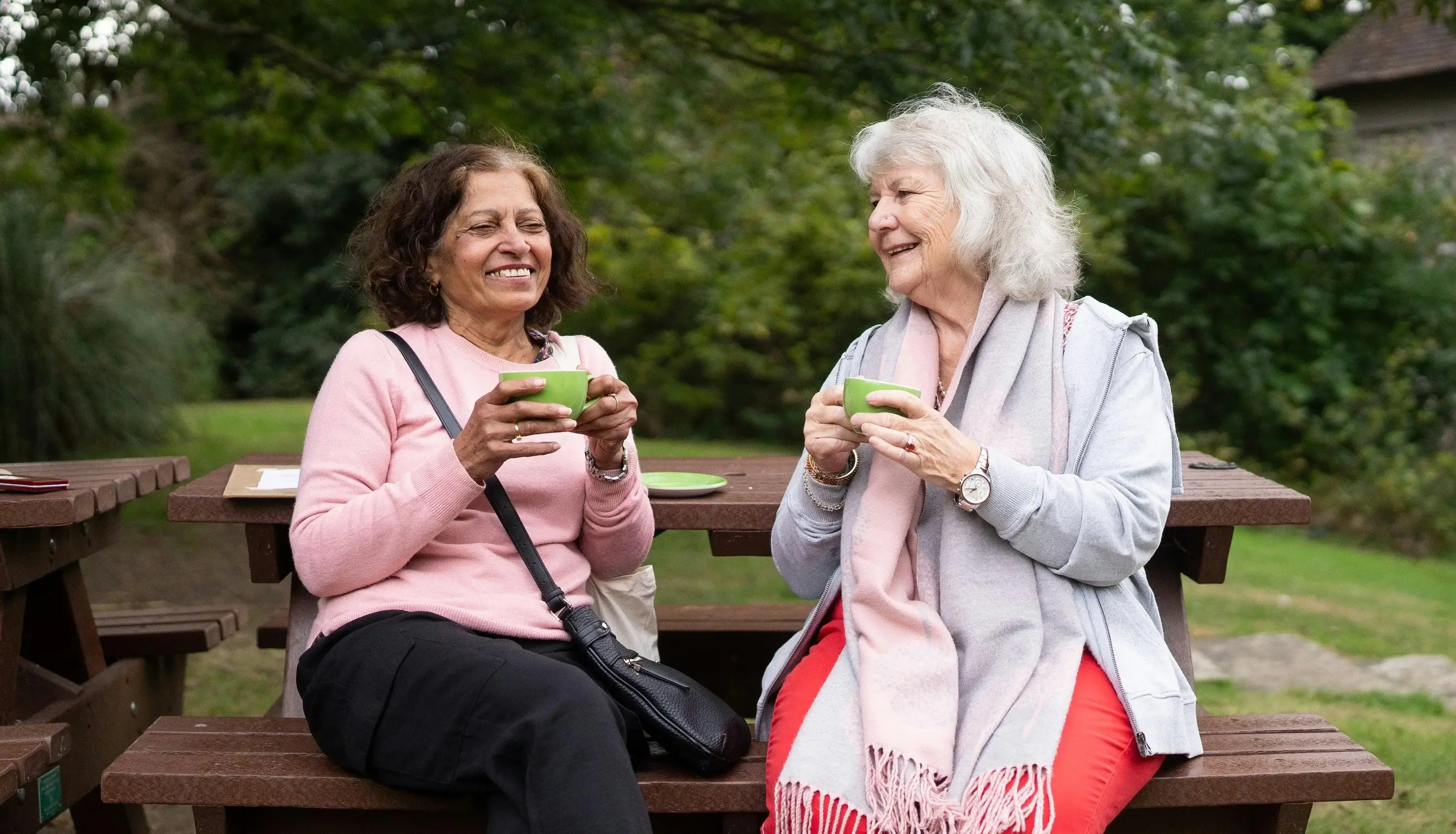 Two older women sitting at a picnic table having tea. 