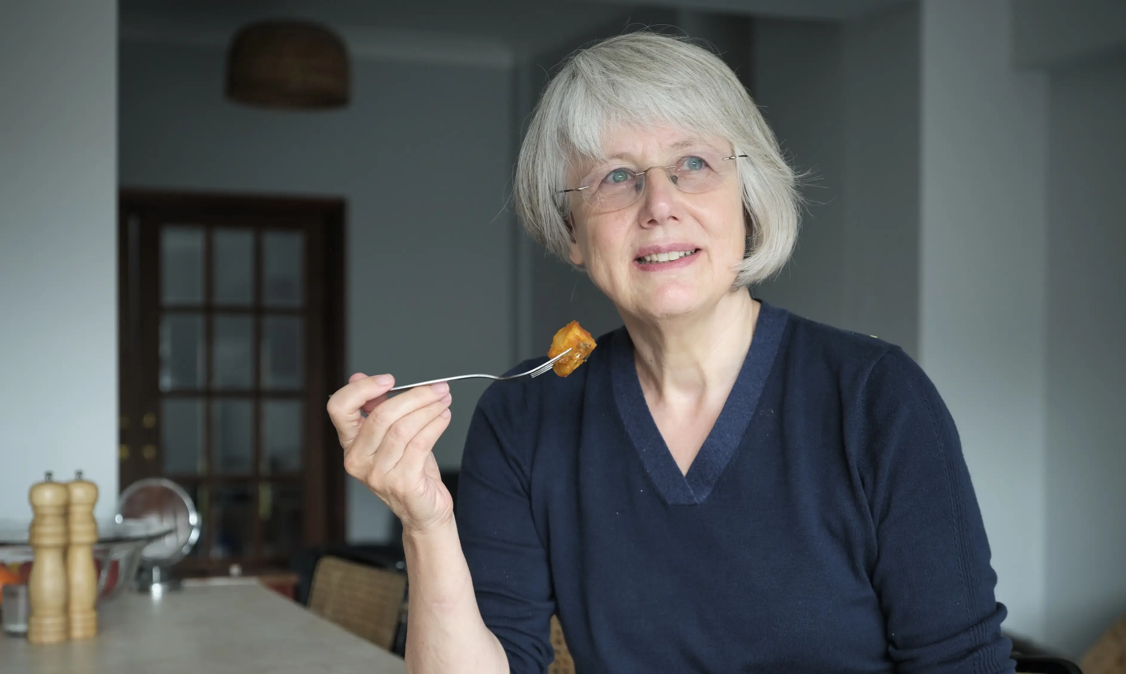A older woman wearing glasses, sitting at her dining table in front of a plate of food. She's eating a meal. 