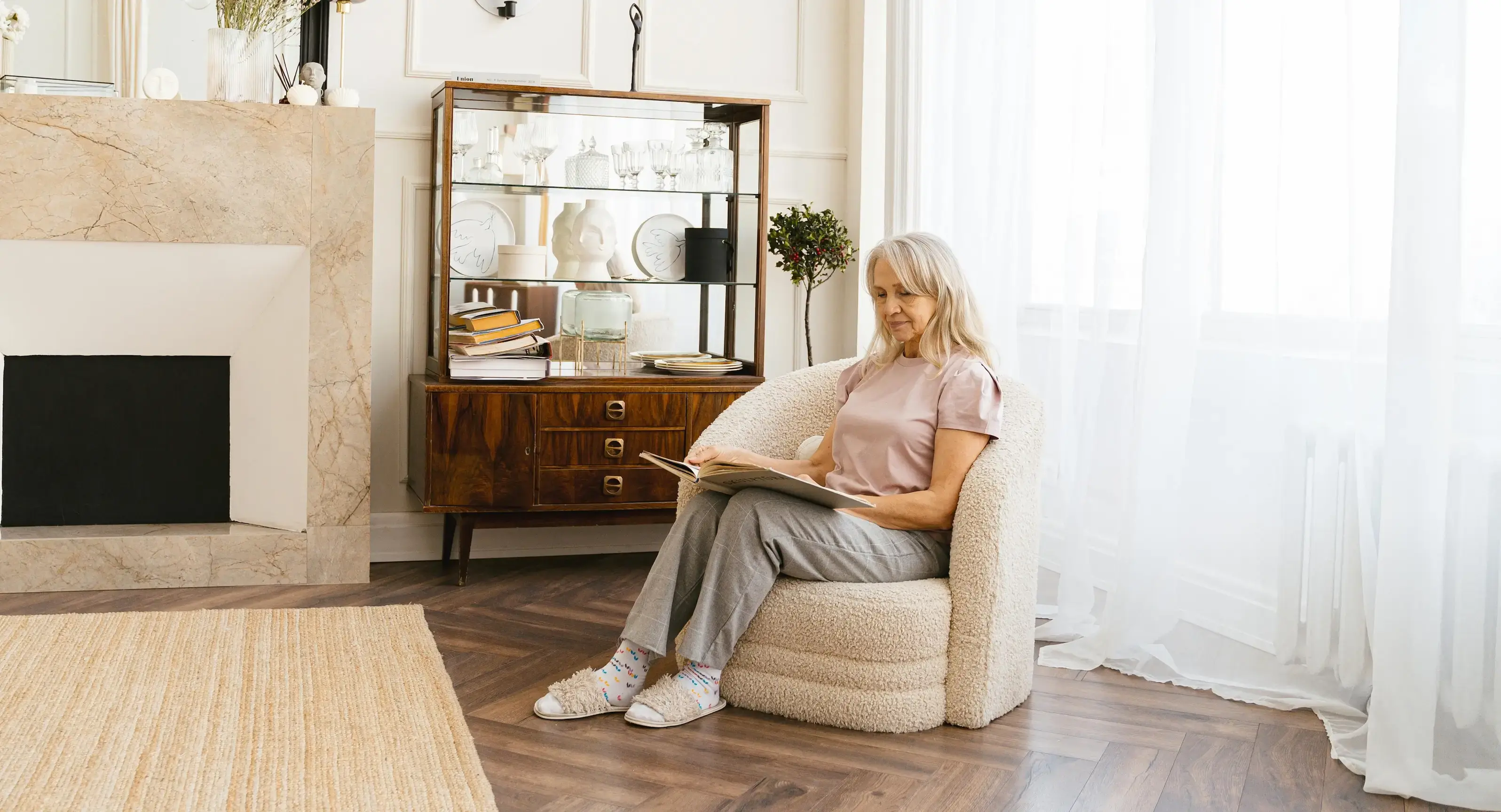 Older woman sitting in an armchair in a well-lit living room, reading a book