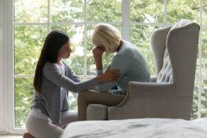 Near panoramic window crying elderly mom sit on armchair, near sit her grown up daughter comforting her in difficult life period give support, share pain at divorce, showing attention and care concept